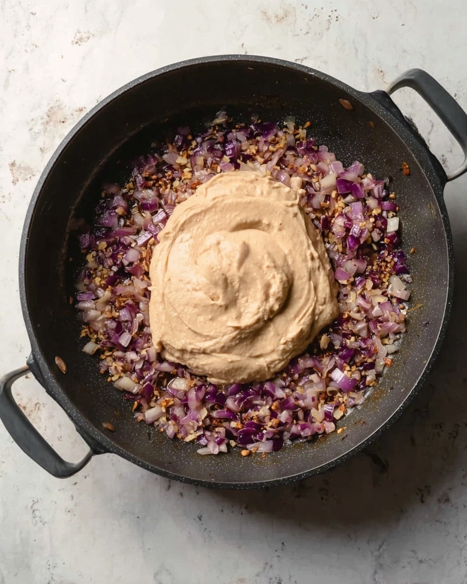 A round dark cooking pan with two handles sits on a white marbled surface, filled with cooked chopped onions and garlic scattered evenly around the pan bottom. In the center, there's a large dollop of light beige creamy sauce with a smooth, slightly textured surface. The edges of the pan show bits of caramelized onion in shades of purple and golden brown. photo taken with an iphone --ar 4:5 --v 7