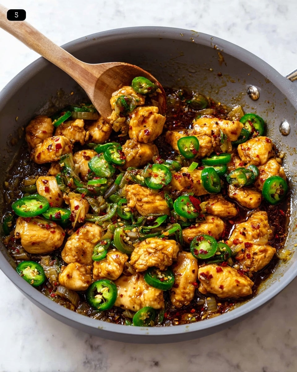 A gray pan filled with several small, golden-brown cooked chicken pieces mixed with bright green sliced jalapeños and bits of cooked onion. The chicken pieces have a slightly crispy texture with a shiny coating of dark brown sauce scattered lightly with red chili flakes. A wooden spoon is stirring the mixture, lifting some chicken pieces and jalapeños from the middle, creating a dynamic and fresh look. The pan sits on a white marbled surface. Photo taken with an iphone --ar 4:5 --v 7