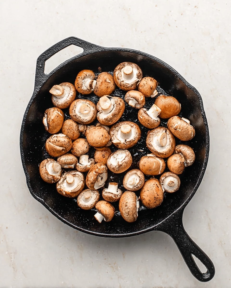 A black cast iron pan is filled with one layer of whole and halved brown mushrooms with white stems facing different directions, lying on a white marbled surface. The mushrooms have a smooth texture and are arranged loosely to cover the pan's surface. The pan handle extends to the right side, and the pan has a coated dark texture that contrasts with the light mushrooms and background. photo taken with an iphone --ar 4:5 --v 7