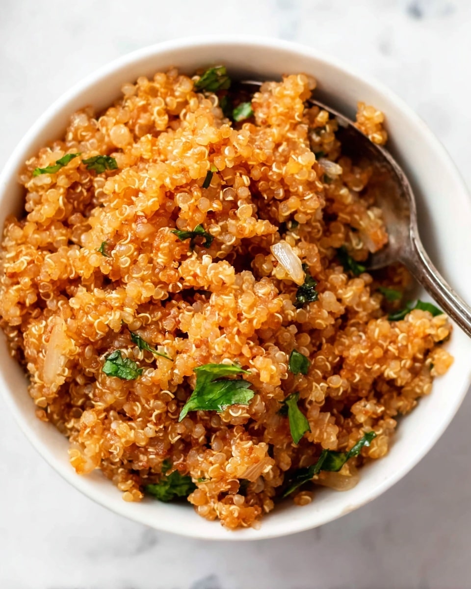 A close-up view of a white bowl filled with cooked quinoa mixed with small pieces of light brown onions and green herbs scattered throughout. The quinoa grains are light orange-brown and fluffy with a slightly wet texture. A silver spoon is partially placed inside the bowl on the right side, with some quinoa resting on it. The bowl sits on a white marbled surface. photo taken with an iphone --ar 4:5 --v 7