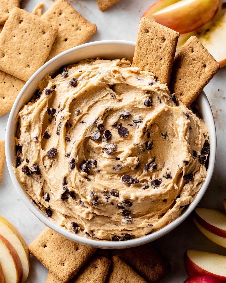 A white bowl filled with thick, creamy cookie dough mixed with small dark chocolate chips, showing textured swirls on the surface. Two rectangular light brown graham crackers are placed sticking out from the dough on the right side. Around the bowl, light brown graham crackers are spread across a white marbled surface, and some slices of red and yellow apples are visible on the lower right side of the image. The overall scene has warm, inviting tones. photo taken with an iphone --ar 4:5 --v 7
