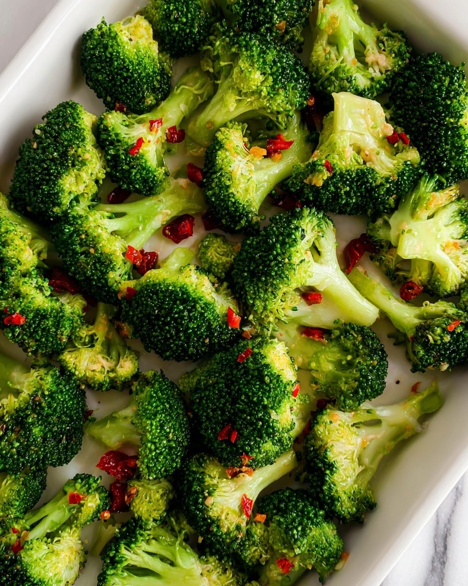 This image shows a white tray filled with many pieces of bright green broccoli florets spread out evenly. Among the broccoli pieces, there are small bits of red pepper scattered throughout, adding a pop of red color. The broccoli has a fresh and slightly shiny texture, suggesting it might be cooked or lightly dressed. The tray sits on a surface with a white marbled texture. The photo is close-up and well-lit, capturing the fresh and colorful nature of the broccoli and red pepper pieces. photo taken with an iphone --ar 4:5 --v 7
