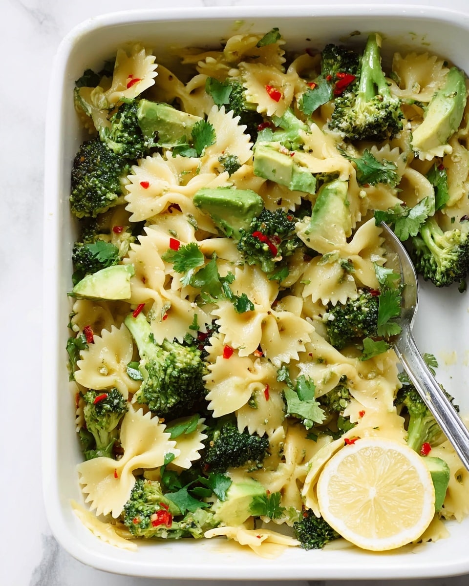 The image shows a white rectangular dish filled with a pasta salad, mainly made of bow-tie pasta and cooked broccoli florets. The pasta is light yellow in color with smooth and slightly curled edges, mixed evenly with small, bright green broccoli pieces with a lightly roasted texture. There are also chunks of avocado scattered throughout, providing a fresh and creamy green contrast. Small bits of red chili pepper and fresh green herbs like cilantro add color pops and freshness. On the right edge of the dish, there is a silver fork partially submerged in the salad, and a half slice of lemon rests close to the corner on a white marbled surface. The overall presentation looks fresh and healthy. photo taken with an iphone --ar 4:5 --v 7