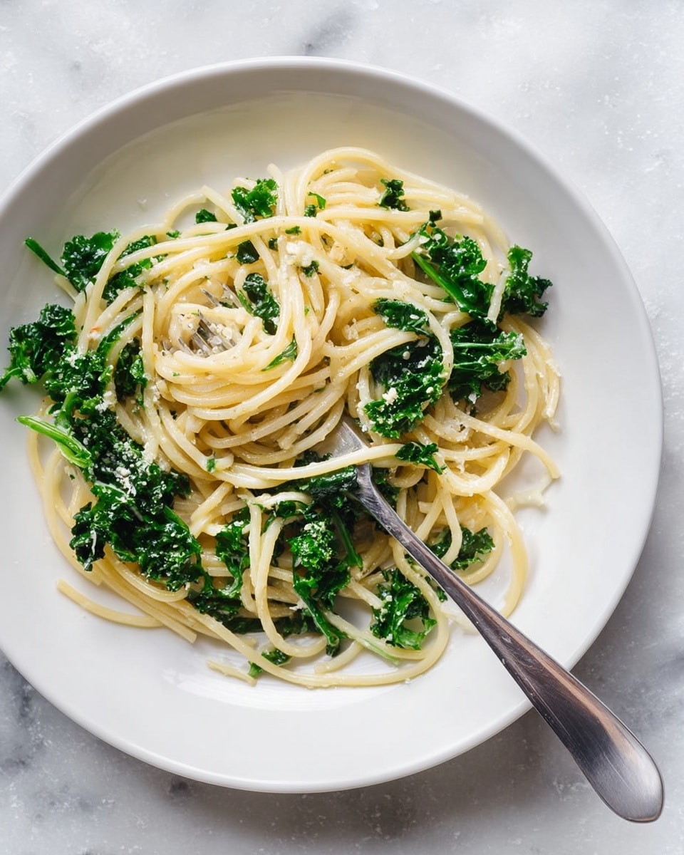 A white plate on a white marbled surface holds a simple pasta dish with spaghetti as the first layer, pale yellow and soft with a smooth texture. Mixed throughout are bright green leafy kale pieces that add a fresh, slightly rough texture. A silver fork is placed on the plate, tangled slightly in the spaghetti with its prongs pointing toward the bottom left. The overall look is light, fresh, and simple with a mix of smooth pasta and leafy greens. photo taken with an iphone --ar 4:5 --v 7