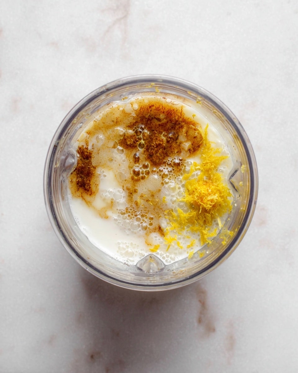 The image shows a clear blender cup placed on a white marbled surface, filled with three visible layers of ingredients. At the bottom, there is a smooth, creamy white liquid base, likely milk or yogurt. On the right side, there is a small pile of bright yellow zest, adding a textured and fresh look. The remaining top layer is a mix of light and dark brown syrup or spices that blend partially into the white liquid below, giving a bubbly and slightly frothy appearance. The overall look is raw and fresh with different textures visible through the clear cup rim. photo taken with an iphone --ar 4:5 --v 7