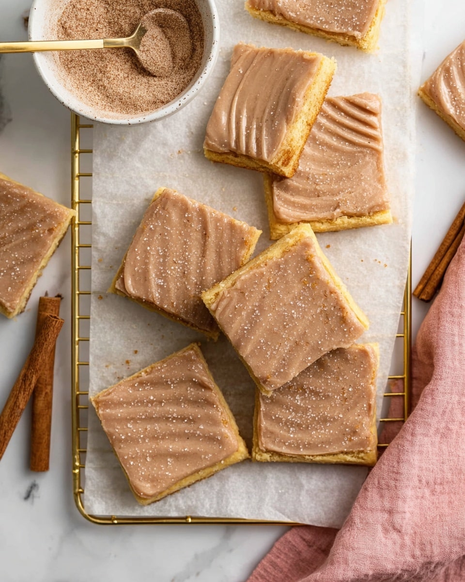 A group of square pastries with a thick light brown frosting layer on top, each pastry showing a golden yellow crust with a fork-pressed edge detail and some parts of the crust slightly exposed under the frosting. The frosting has a smooth texture with a light sprinkling of sugar crystals that adds a subtle sparkle. The pastries are placed on a gold wire rack lined with white parchment paper over a white marbled surface. A white bowl filled with a fine powdery cinnamon-sugar mix is nearby, with a gold spoon resting inside. Cinnamon sticks and a soft pink cloth add warm accents to the scene. Photo taken with an iphone --ar 4:5 --v 7