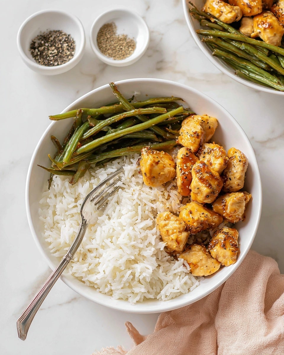 The image shows a white bowl filled with a meal of three layers: the first layer is white fluffy rice filling half of the bowl, the second layer is a portion of cooked green beans with a slightly shiny texture placed on the top right, and the third layer at the bottom right is bite-sized pieces of golden-brown chicken with a glossy sauce and sprinkle of black pepper. A silver fork rests on the rice towards the center of the bowl. In the background, there is a smaller white bowl with black pepper and salt on a white marbled surface, accompanied by another bowl with the same meal partially visible. A beige cloth is placed near the bottom right corner. photo taken with an iphone --ar 4:5 --v 7
