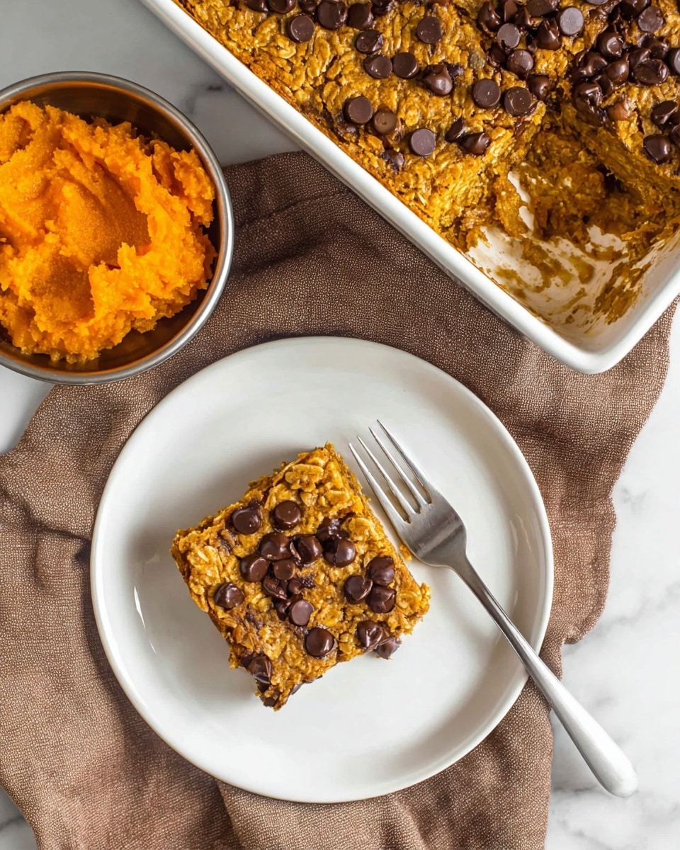 A single square piece of chocolate chip oatmeal bar sits in the center of a white plate, showing a golden-brown textured top with melted dark chocolate chips scattered on it. The bar’s surface looks moist and slightly rough with visible oatmeal bits. A silver fork rests on the plate next to the bar. Above the plate is a white baking dish filled with the same chocolate chip oatmeal bars, its surface uneven with more dark chocolate chips on top. To the left of the scene is a small metal container with bright orange mashed pumpkin inside. All items are placed on a soft brown cloth that covers a white marbled surface. photo taken with an iphone --ar 4:5 --v 7