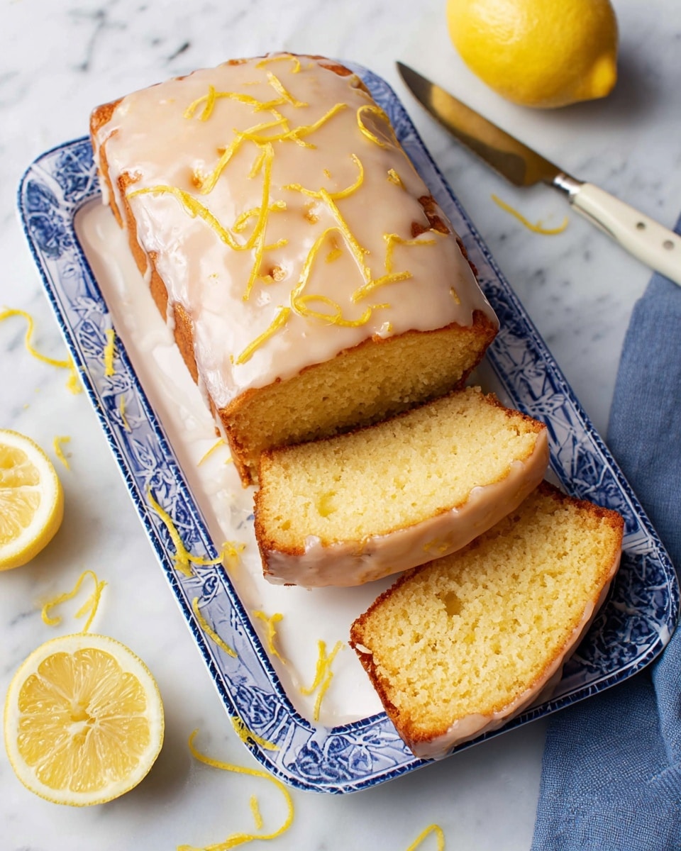 A white plate with blue patterns holds a rectangular lemon loaf cake with a light golden brown crust. The cake has three visible slices showing a soft, moist yellow crumb inside. The top is covered with a smooth, light beige glaze, drizzled evenly, with small bright yellow lemon zest pieces scattered on top and around the plate. Two lemon halves are placed nearby on a white marbled surface, adding bright yellow color. Part of a small knife with a white handle is visible at the side, along with a blue cloth napkin in the corner. Photo taken with an iphone --ar 4:5 --v 7
