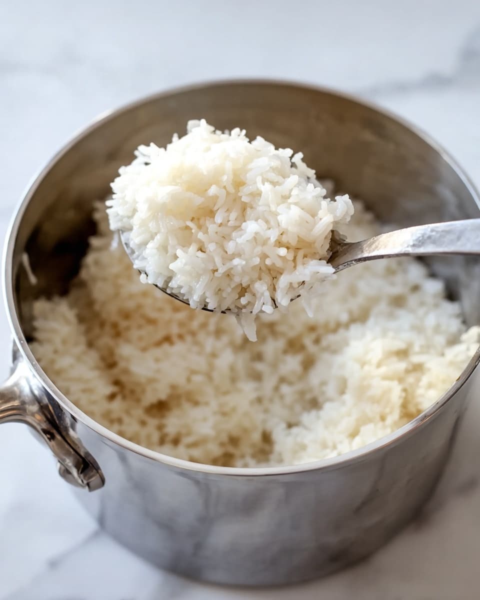 The image shows a shiny silver pot filled with fluffy white cooked rice. A silver spoon lifts a scoop of soft, sticky white rice from the pot, with the rice grains clearly visible and lightly clumped together. The pot is placed on a white marbled surface, and the soft lighting highlights the slight glossiness of the rice. The simple scene focuses on the texture and whiteness of the cooked rice against the metallic pot and clean background. Photo taken with an iphone --ar 4:5 --v 7