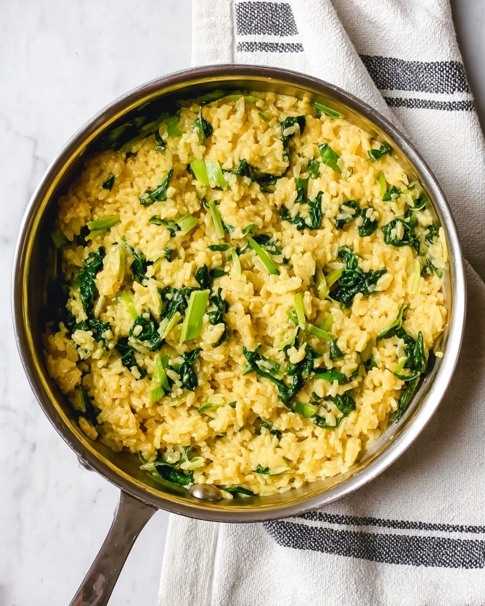 A close-up view of a stainless steel pan filled with a creamy yellow rice dish mixed with bright green leafy vegetables and chopped stalks, evenly spread inside the pan. The rice looks soft and thick, with the green vegetable pieces scattered throughout the creamy texture, creating a mix of green and yellow colors. The pan is placed on a white cloth with thin black stripes, on a white marbled surface. photo taken with an iphone --ar 4:5 --v 7