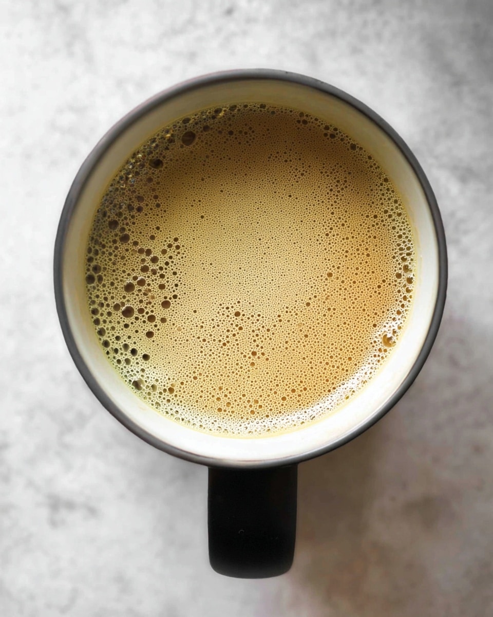 A top view of a single cup filled with light brown coffee with a layer of small bubbles on the surface, inside a white cup that has a smooth black handle seen at the bottom center, all placed on a white marbled textured surface photo taken with an iphone --ar 4:5 --v 7