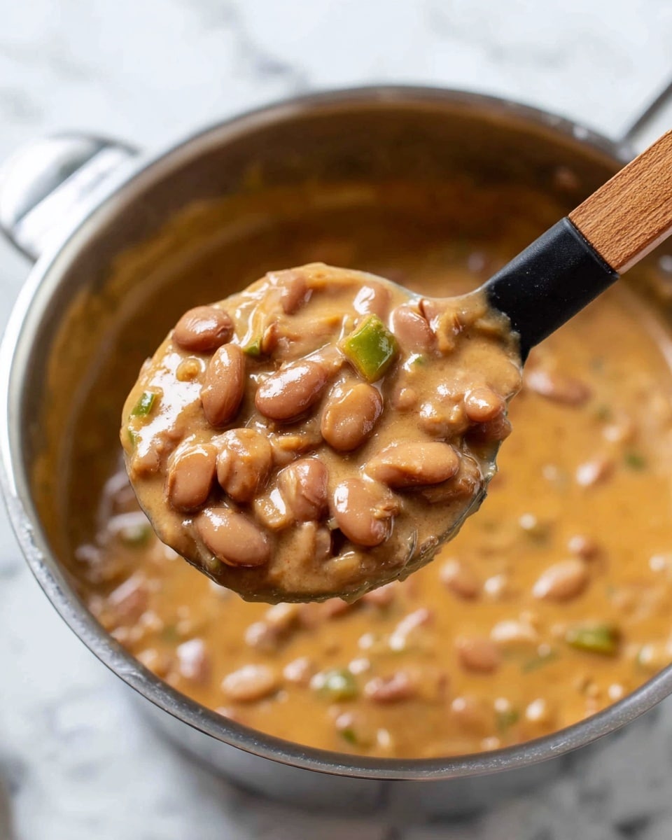 The image shows a close-up of a pot filled with creamy beans stew. The stew has a thick, smooth texture mixed with whole brown beans and small pieces of green pepper, all coated in a light brown sauce. A black spoon with a wooden handle holds a scoop of the beans stew above the pot, showing the chunky and creamy consistency clearly. The pot is silver, and the background has a white marbled texture. photo taken with an iphone --ar 4:5 --v 7