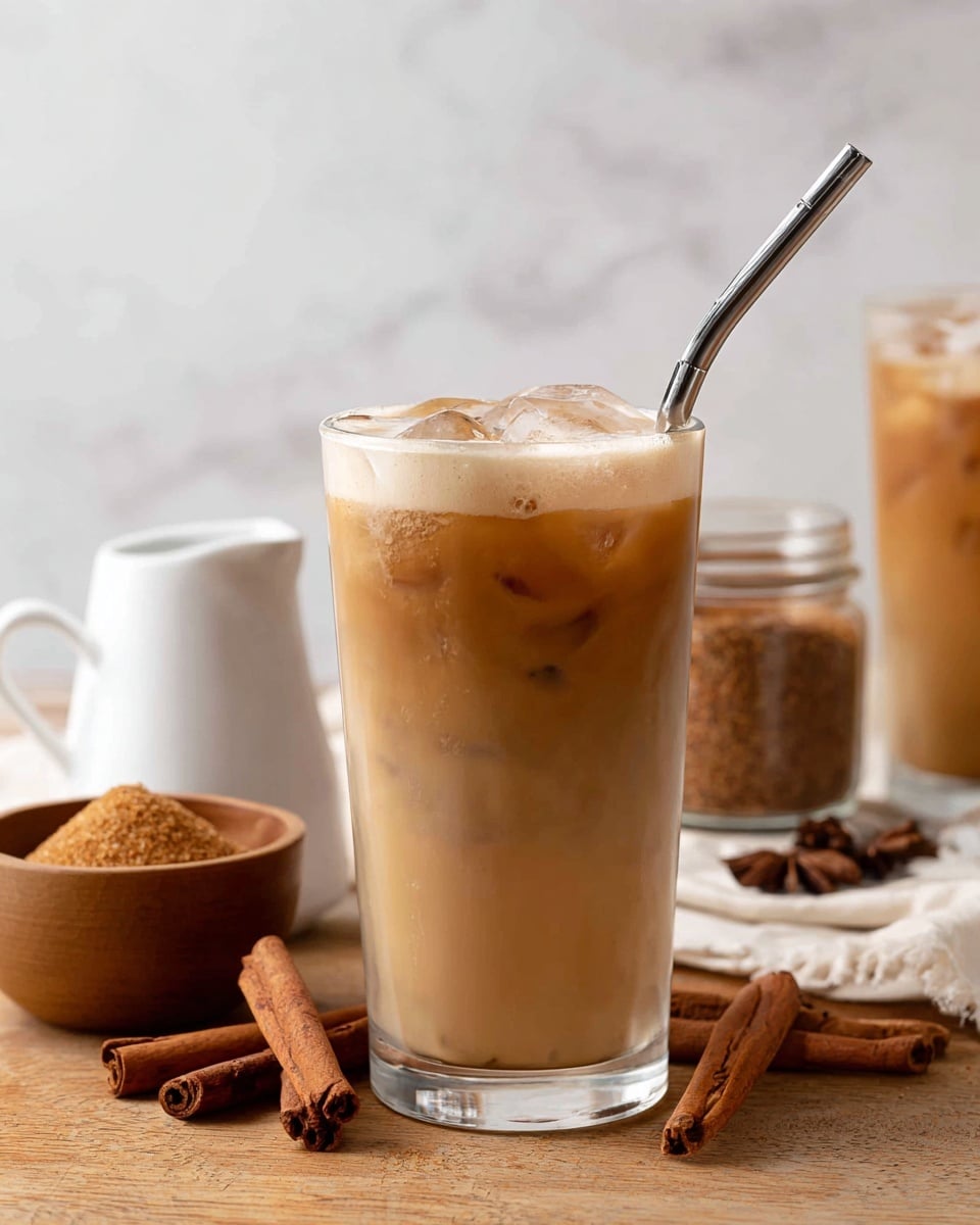 A tall clear glass filled with a light brown iced coffee layered with ice cubes inside, topped with a thick frothy cream layer, and a shiny metal straw on the right side; in the background, another glass of iced coffee, a small white creamer pitcher, a wooden bowl full of brown sugar, a glass jar, and cinnamon sticks lay on a wooden surface, all placed on a white marbled texture background. photo taken with an iphone --ar 4:5 --v 7