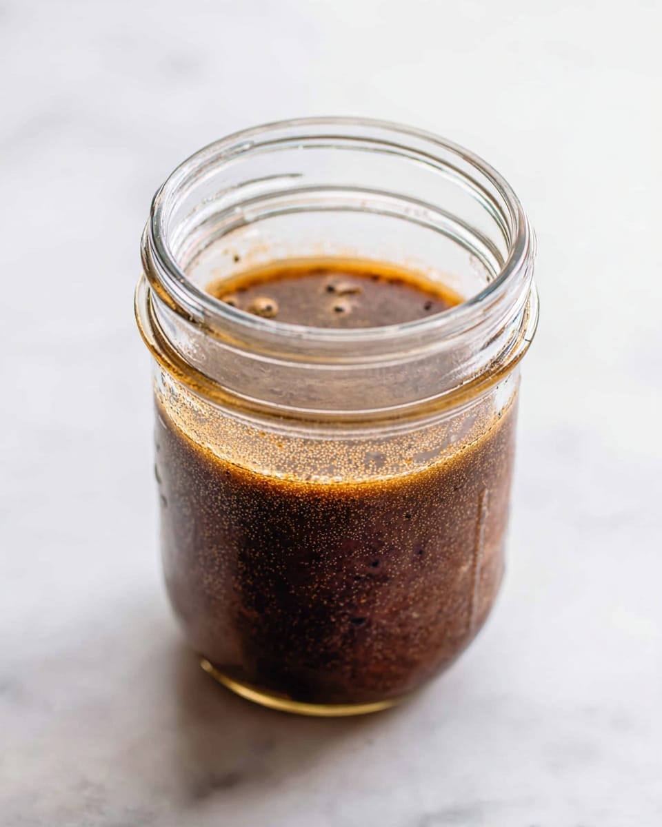 A clear glass mason jar filled about halfway with a dark brown, slightly grainy liquid. The liquid has small black specks and some oil droplets floating on the surface, giving it a textured look. The jar sits on a smooth white marbled surface, and the background is softly lit to create a clean and bright setting. photo taken with an iphone --ar 4:5 --v 7