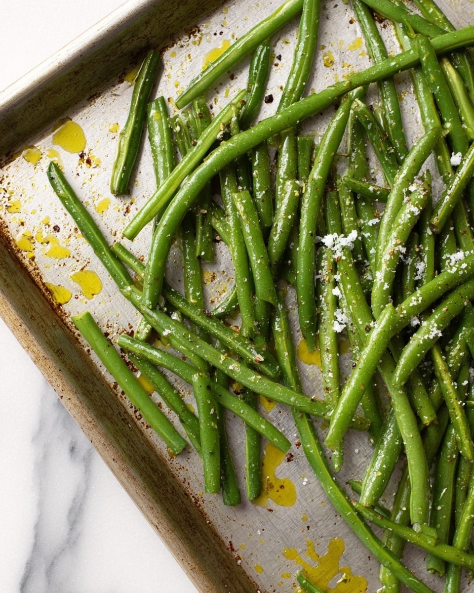 The image shows a close-up of fresh green beans spread out on a lightly used metal baking tray. The green beans are bright green, some longer and some shorter, with a slight shine from oil or dressing, scattered unevenly across the tray. Small white granules, likely salt and some powdery seasoning, are sprinkled over the beans. There are small drops of yellow oil and black pepper flakes visibly resting on the tray’s surface, which is scratched and shows signs of use. The background under the tray is a white marbled texture. photo taken with an iphone --ar 4:5 --v 7