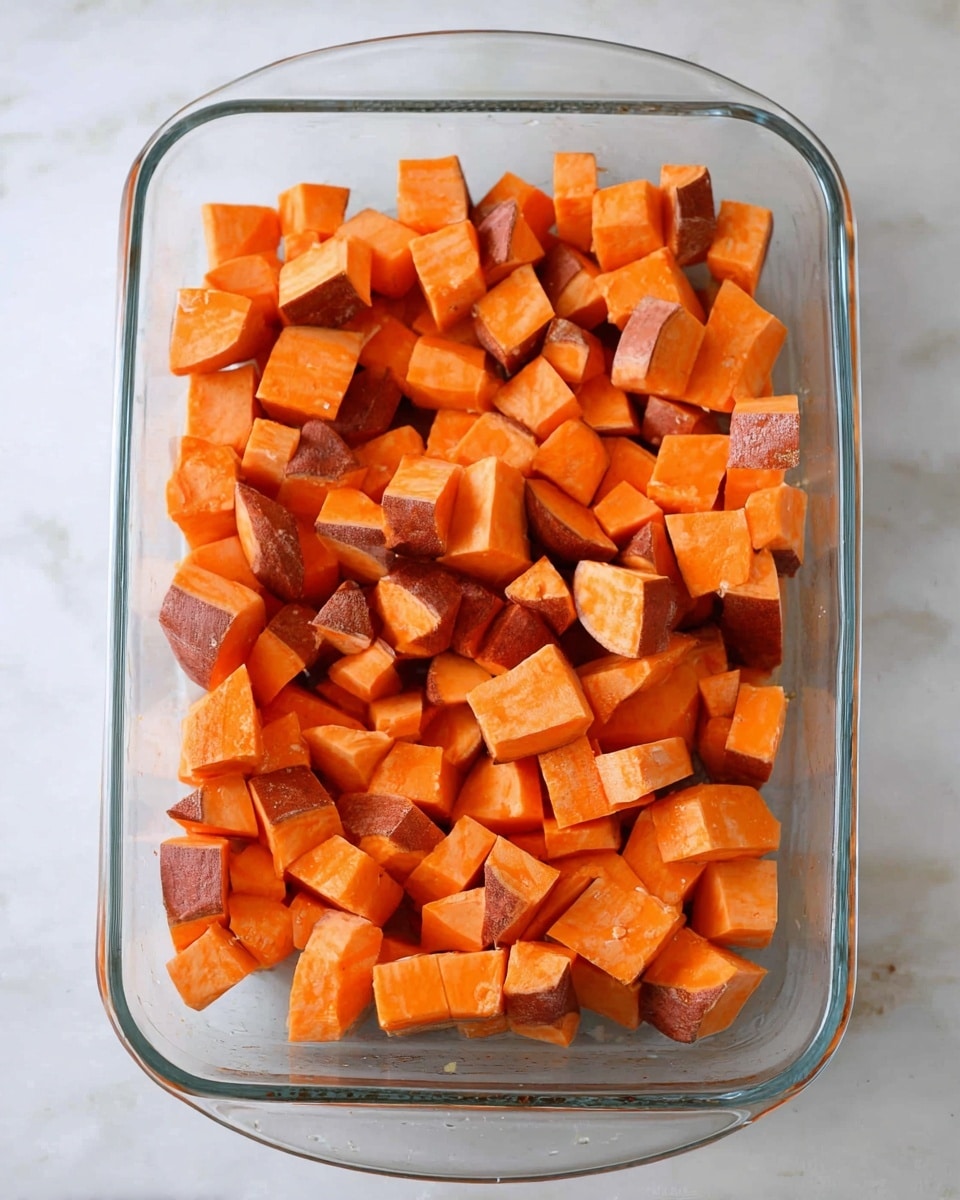 A clear glass baking dish is filled with evenly sized cubes of sweet potatoes with orange flesh and reddish-brown skin, filling the dish completely. The baking dish rests on a white marbled surface with soft, diffused lighting highlighting the bright orange color and slight texture of the sweet potatoes. Photo taken with an iphone --ar 4:5 --v 7