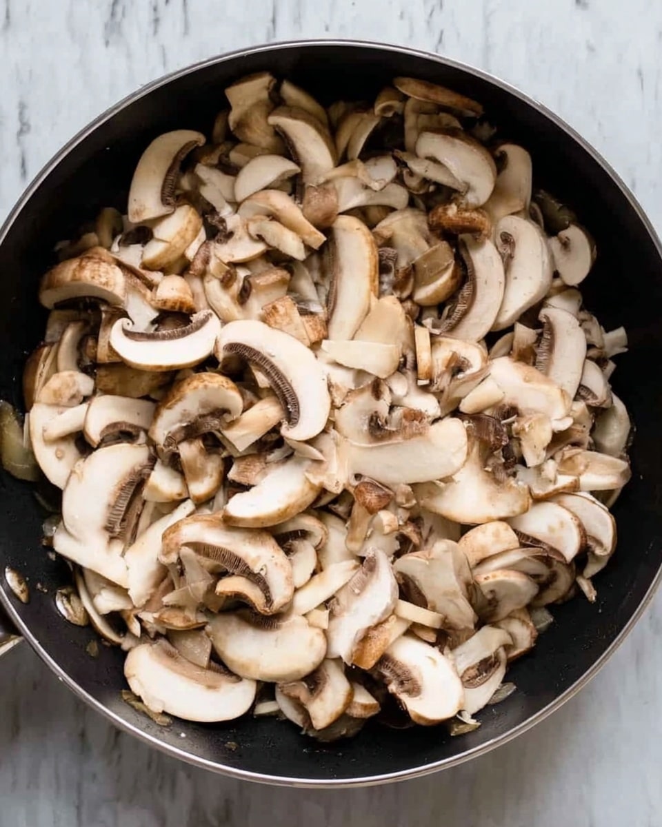 An overhead view shows a black frying pan filled with sliced white mushrooms. The mushrooms vary in size and shape, with some showing the white inside and brown gills, while others show the light brown outside. The mushrooms fill the whole pan, and their smooth, slightly shiny texture is clear. The pan sits on a white marbled surface with subtle gray veins. Photo taken with an iphone --ar 4:5 --v 7