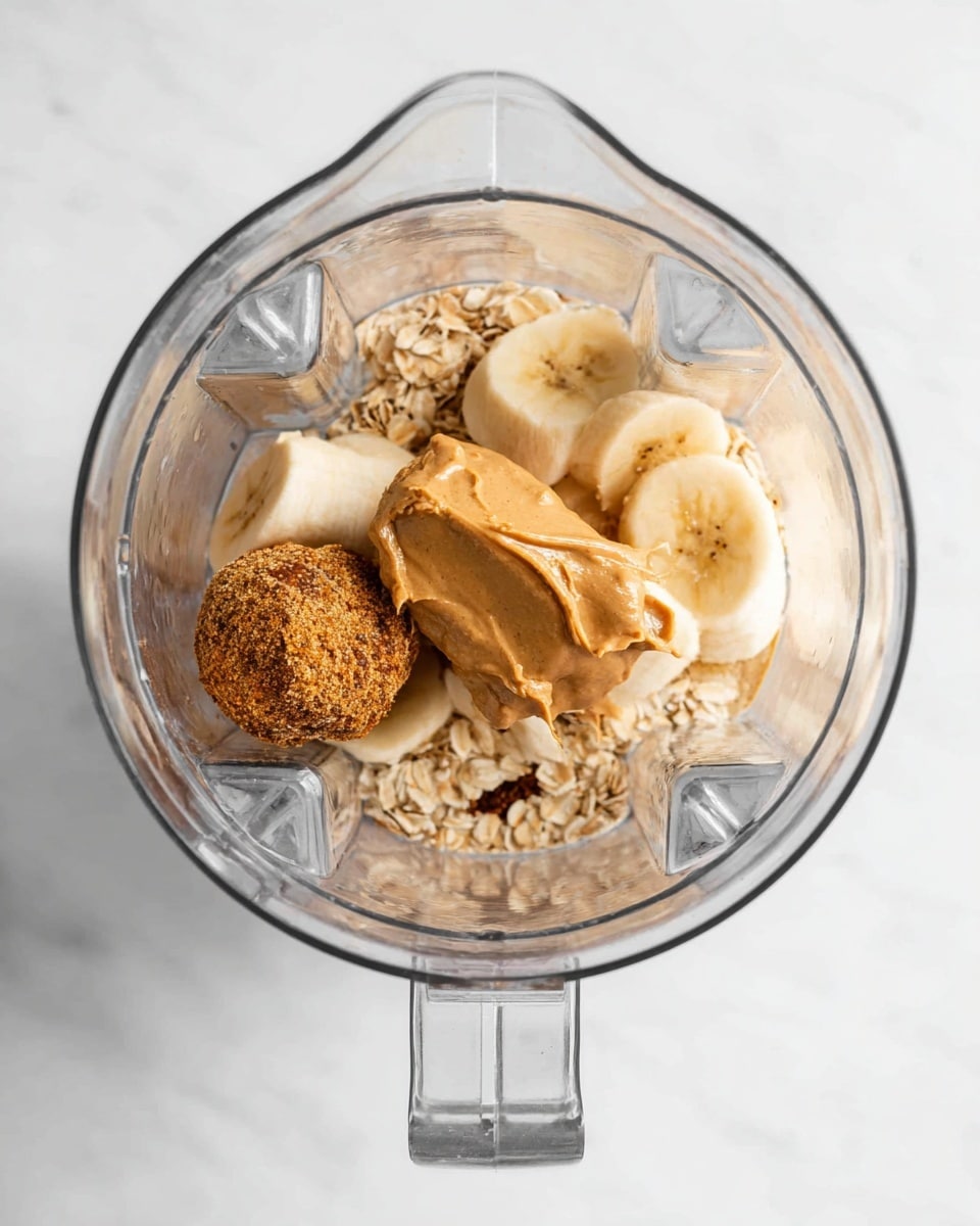 A top view shows a blender jar with different ingredients inside, layered loosely. The first layer is a light brown spoon of peanut butter with a creamy but textured appearance on the right side. Next to it, there are several thick slices of banana, pale yellow with a soft texture. Scattered around are few light tan oats. At the bottom left is a round brown ball, looking like a date or energy bite, with a rough textured surface. The base of the jar and the little dark powder particles can be seen beneath the ingredients. The background is a white marbled texture. Photo taken with an iphone --ar 4:5 --v 7