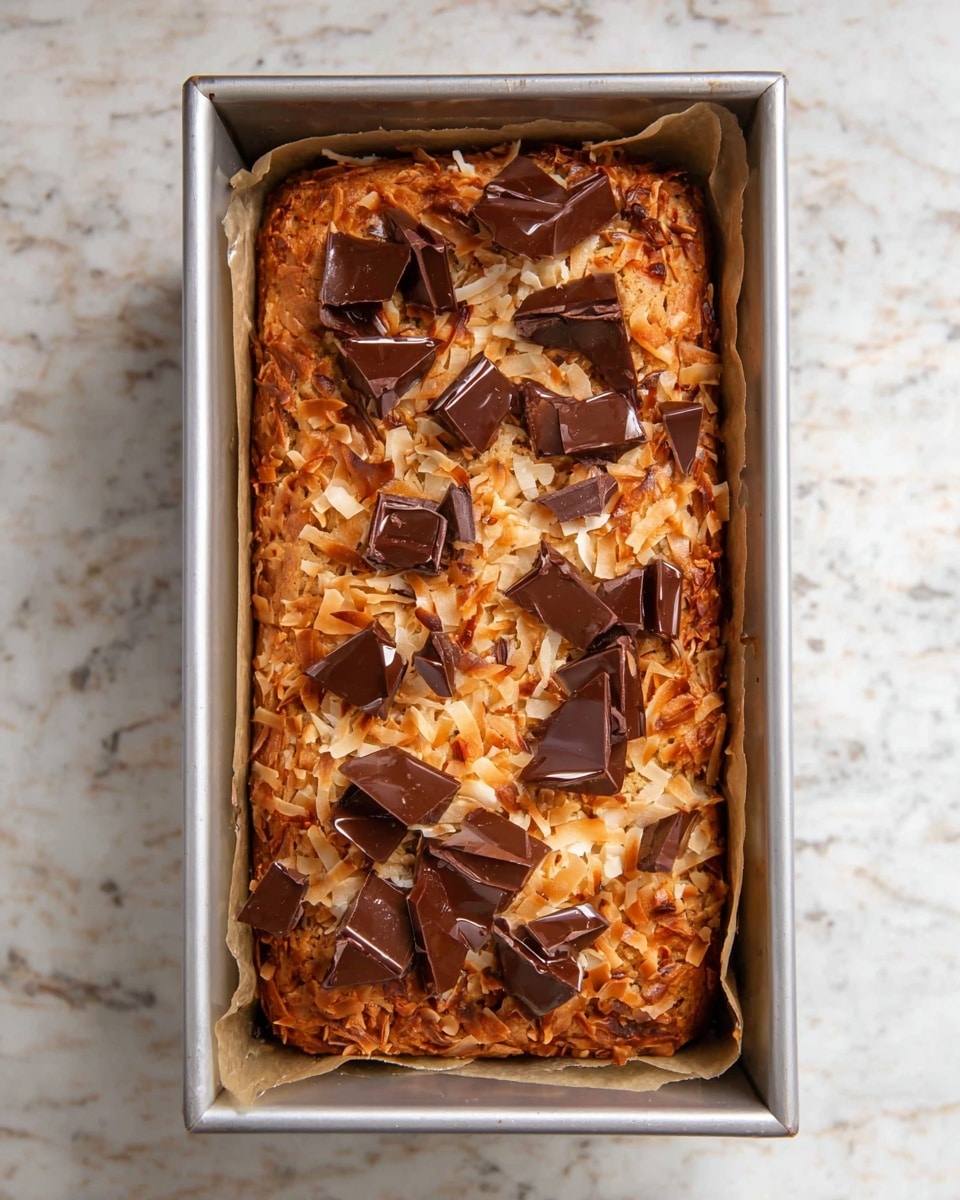 The image shows a loaf in a metal baking pan lined with parchment paper. The top layer has golden brown toasted shredded coconut spread evenly, with large, glossy dark brown chocolate pieces scattered across it. The coconut has a rough, crispy texture with some darker toasted spots, and the chocolate pieces appear slightly melted. The pan is placed on a white marbled surface that looks clean and bright. photo taken with an iphone --ar 4:5 --v 7
