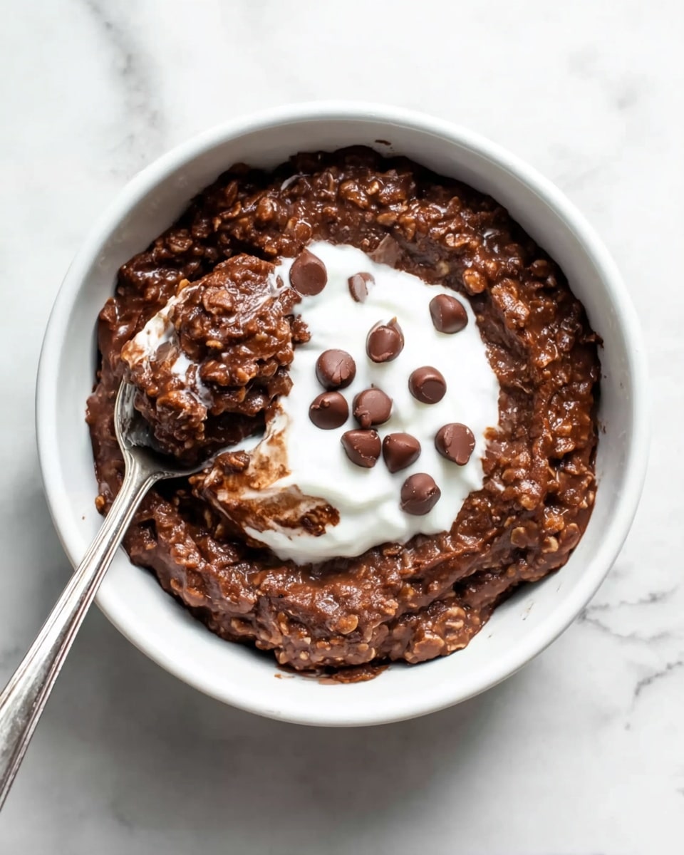 The image shows a white bowl filled with thick, dark brown chocolate oatmeal with a slightly chunky texture. In the center of the oatmeal is a dollop of smooth white cream or yogurt, topped with several round, glossy chocolate chips scattered across the cream and oatmeal. A silver spoon is placed inside the bowl on the left side, partly submerged in the oatmeal, with some chocolate oatmeal clinging to the spoon’s edge. The bowl sits on a white marbled surface. photo taken with an iphone --ar 4:5 --v 7