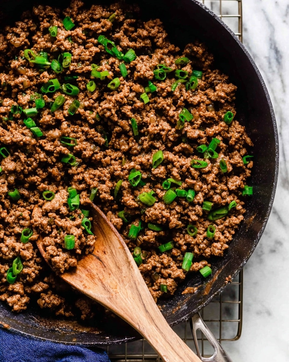 A close-up top view of a black skillet filled with cooked ground meat mixed with small pieces of green onions scattered evenly on top. A wooden spoon is scooping some of the meat from the right side, blending smoothly into the textured brown meat layers. The skillet rests on a wire rack above a white marbled surface. photo taken with an iphone --ar 4:5 --v 7