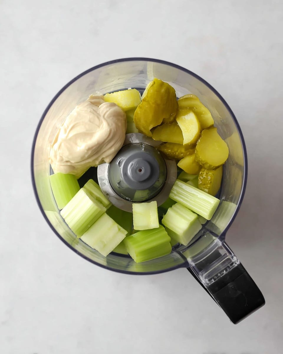The image shows a clear food processor bowl with four main items inside, placed on a white marbled surface. At the bottom layer, there are three light green pieces of celery with a crunchy texture, placed around the bowl's center. On top of the celery, there are three yellowish-green chunks of pickles, glossy and slightly wet in appearance, positioned evenly. To the left side of the bowl's center, a dollop of creamy, pale beige mayonnaise rests smoothly with a soft texture. The food processor's metal blade is visible in the center among the ingredients, and a black handle extends to the left outside the bowl's edge. photo taken with an iphone --ar 4:5 --v 7