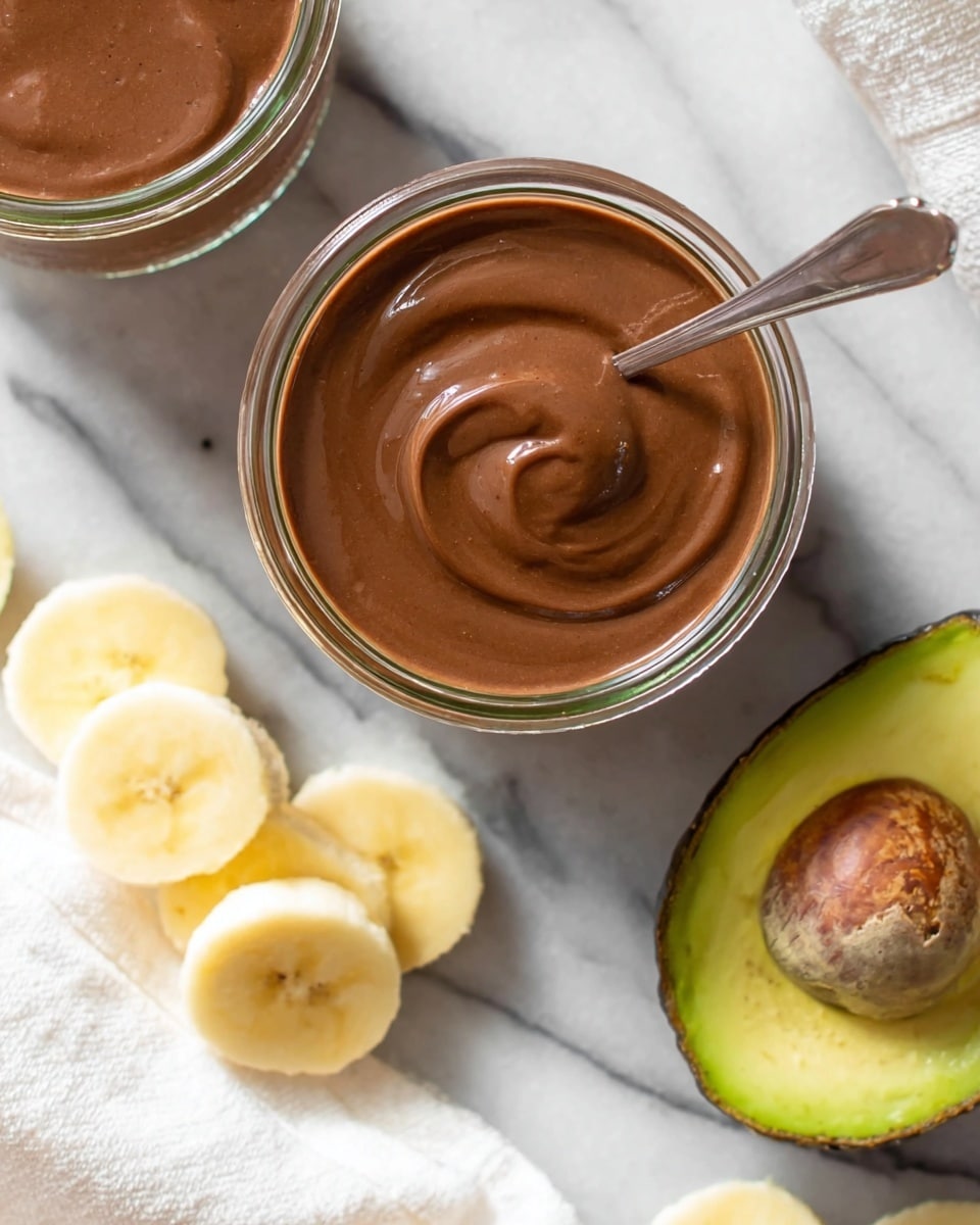 The image shows two clear glass jars filled with smooth, thick brown chocolate sauce, placed on a white marbled surface. One jar is seen from the top, with a silver spoon inside the sauce, creating a small swirl pattern. The other jar is partly visible in the top left. On the right side, an avocado half with green flesh and a brown pit rests on the surface. At the bottom left, several bright yellow banana slices sit on a white cloth, slightly overlapping each other. The scene is bright and clean, focusing on the rich chocolate sauce and fresh fruit elements. photo taken with an iphone --ar 4:5 --v 7