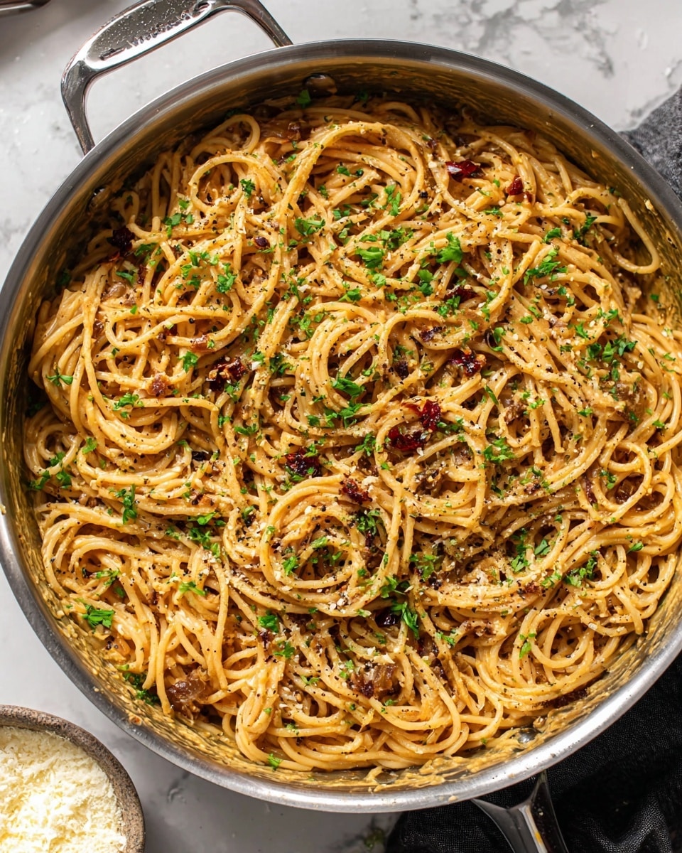 A large metal pan filled with spaghetti pasta mixed in a creamy light brown sauce, with small bits of cooked onions and red chili flakes scattered throughout. The pasta is garnished with chopped green herbs, likely parsley, and sprinkled with grated cheese and black pepper, creating a textured and vibrant look. The pan handle is visible at the top left, and a small bowl with grated cheese is partially seen on a white marbled surface in the background. photo taken with an iphone --ar 4:5 --v 7