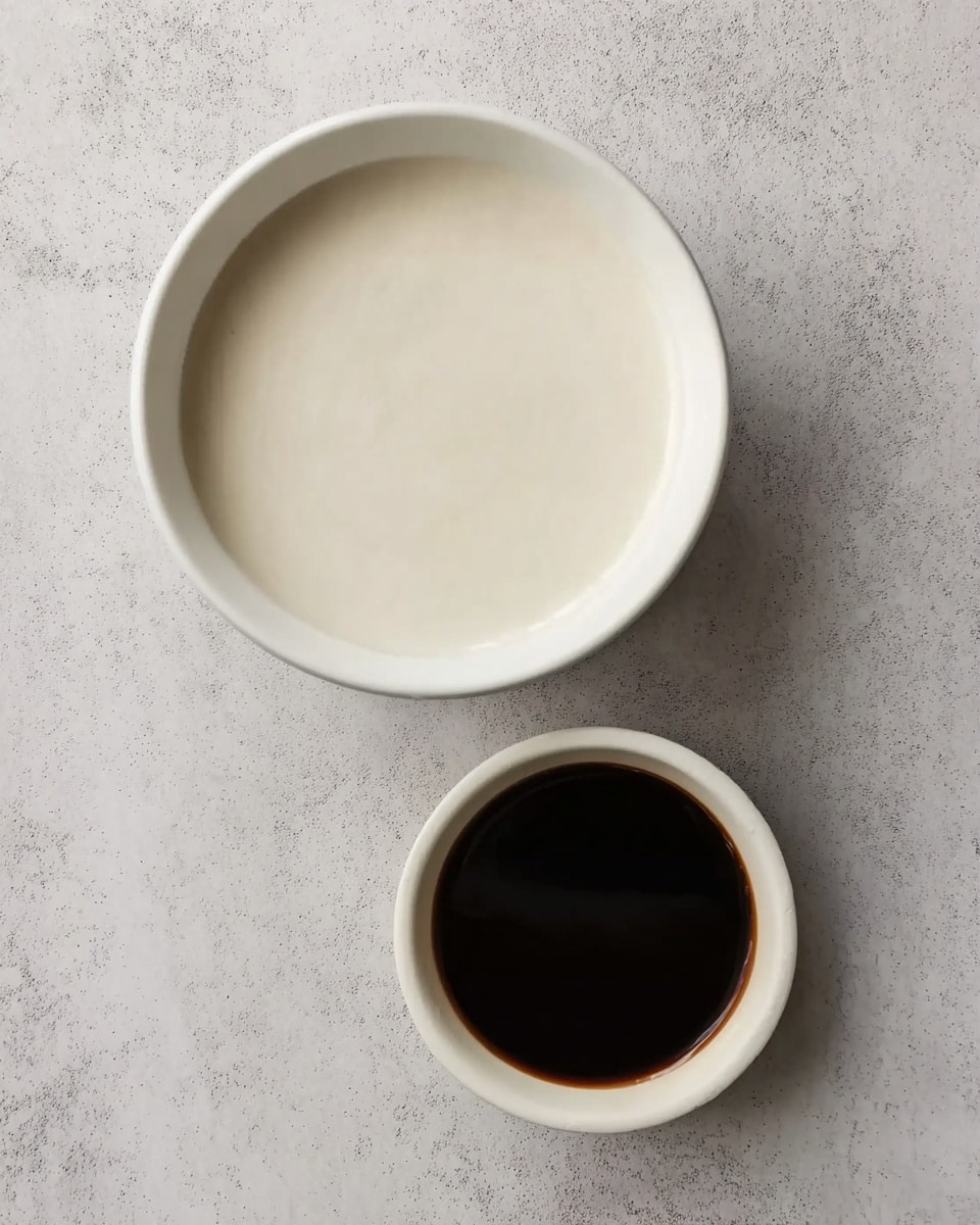 The image shows two white round bowls placed on a white marbled surface. The larger bowl at the top is filled with a smooth, off-white liquid that looks creamy and thick. Below it, the smaller bowl contains a dark brown liquid that is shiny with a smooth texture. Both bowls are simple and clean, with no patterns or decorations. Photo taken with an iphone --ar 4:5 --v 7