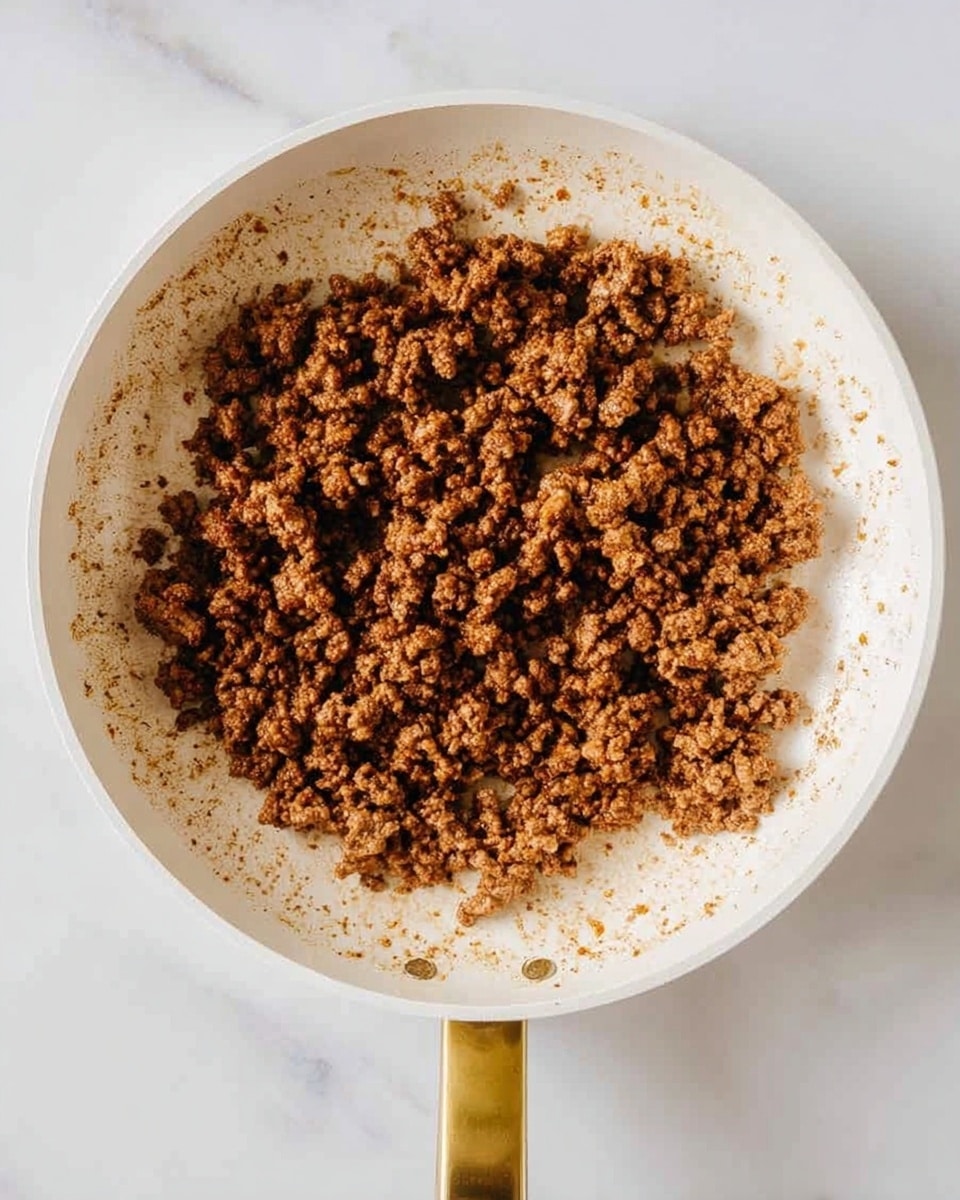 A white frying pan filled with cooked ground meat that is brown in color with a crumbly texture. The pan handle is gold and the cooking surface has some browned bits and spices around the edges. The pan is placed on a white marbled surface. Photo taken with an iphone --ar 4:5 --v 7