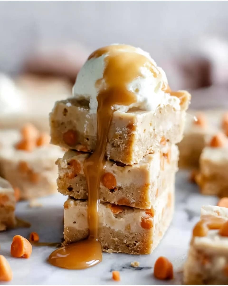 A stack of three light beige bars with small orange dots, likely chips, is placed on a white marbled surface. The bars have a soft, crumbly texture and are neatly square-shaped. On top of the stack, there is a dollop of creamy white ice cream melting slightly, with thick brown caramel sauce dripping down the sides of the bars. In the background, more bars and scattered orange chips are visible but out of focus. The photo has a close-up view, emphasizing the texture and melting sauce, photo taken with an iphone --ar 4:5 --v 7