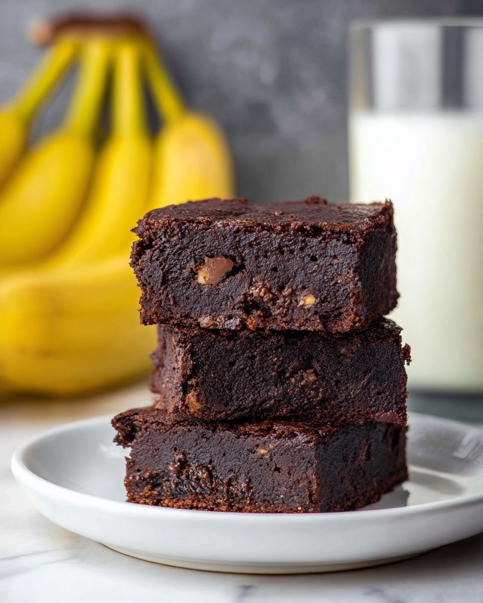 Three dark brown chocolate brownies are stacked on top of each other on a white plate. Each brownie has visible chocolate chunks inside, showing a dense and moist texture. Behind the brownies, there is a clear glass filled with white milk and some yellow bananas slightly out of focus. The surface is white marbled texture. photo taken with an iphone --ar 4:5 --v 7