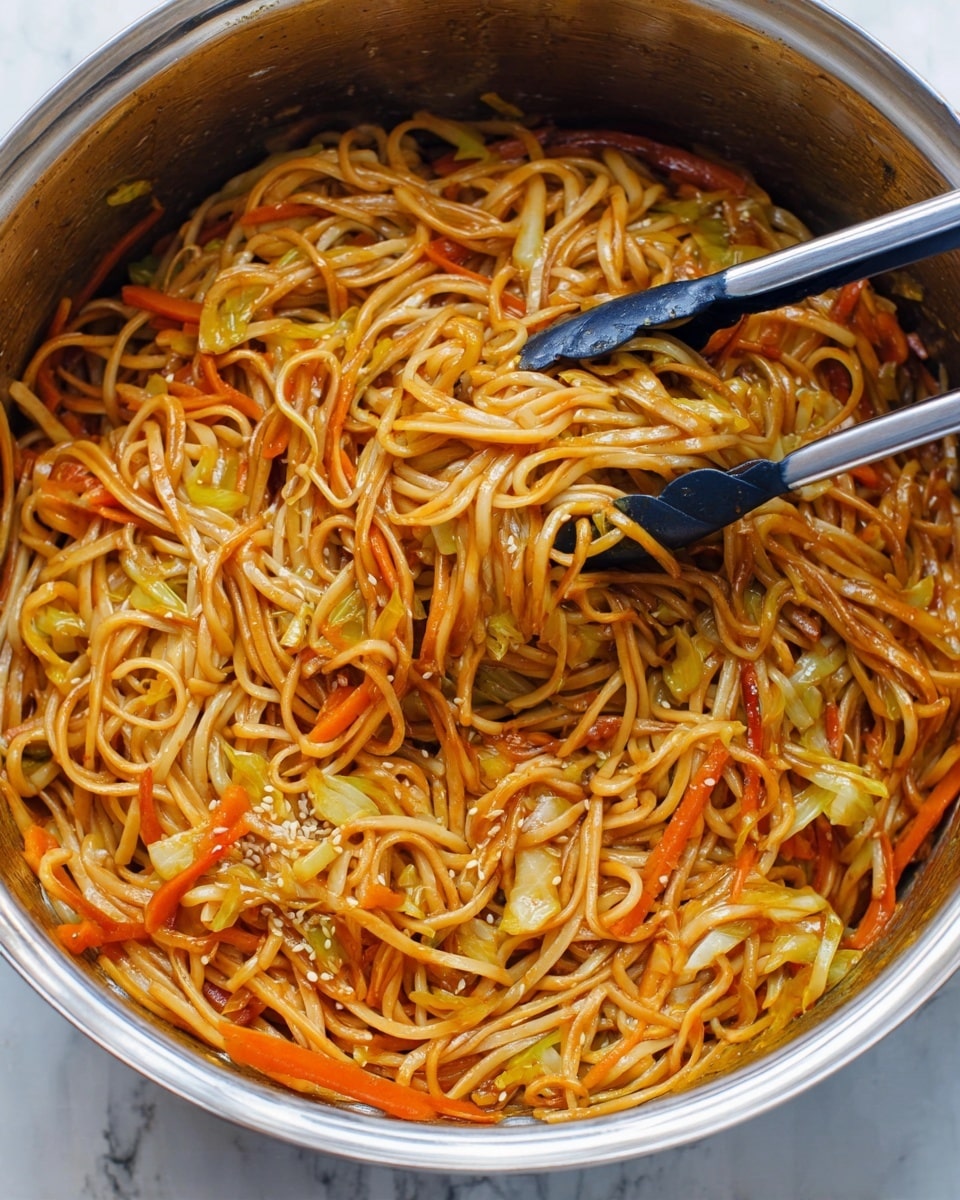 A large metal pot filled with a single layer of cooked noodles mixed with thin slices of orange carrots and pale green cabbage, all coated in a shiny reddish-brown sauce. The noodles are long, tangled, and soft, with sesame seeds lightly sprinkled on top. A pair of black and silver kitchen tongs lifts a small clump of the noodles and vegetables from the pot. The pot sits on a white marbled surface. Photo taken with an iphone --ar 4:5 --v 7