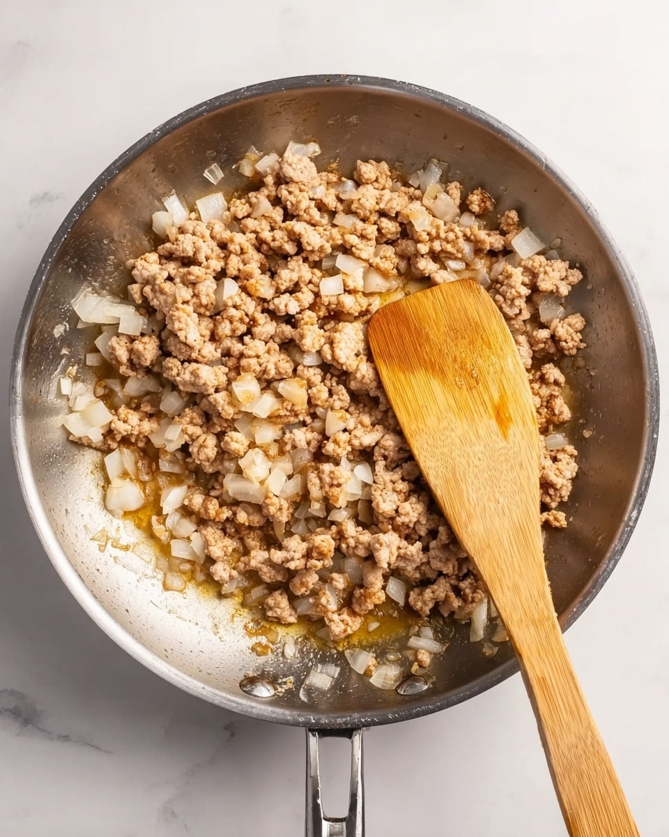 A shiny silver pan contains cooked ground meat mixed with small pieces of white onion. The meat is light brown with some golden spots, and the onions are small, square, and translucent. A wooden spatula with a smooth texture is placed inside the pan, slightly lifting some of the mixture. The pan sits on a white marbled surface that has soft grey veins. The scene is brightly lit, showing the details of the cooked food and the metallic shine of the pan. photo taken with an iphone --ar 4:5 --v 7