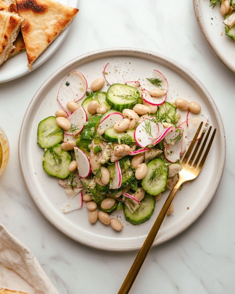 The image shows a white plate filled with a fresh salad made of three main layers: the first layer consists of green cucumber slices with a slightly glossy texture, the second layer is made up of small, smooth white beans scattered evenly, and the third layer includes thin, white radish strips with pink edges mixed with small green herb pieces, possibly dill, sprinkled throughout. The salad is lightly seasoned with visible black pepper specks on top. A gold fork sits on the right side of the plate, resting partially on the salad. The plate is placed on a white marbled surface, with part of a white plate holding triangular toasted flatbread pieces visible in the upper left corner. Photo taken with an iphone --ar 4:5 --v 7