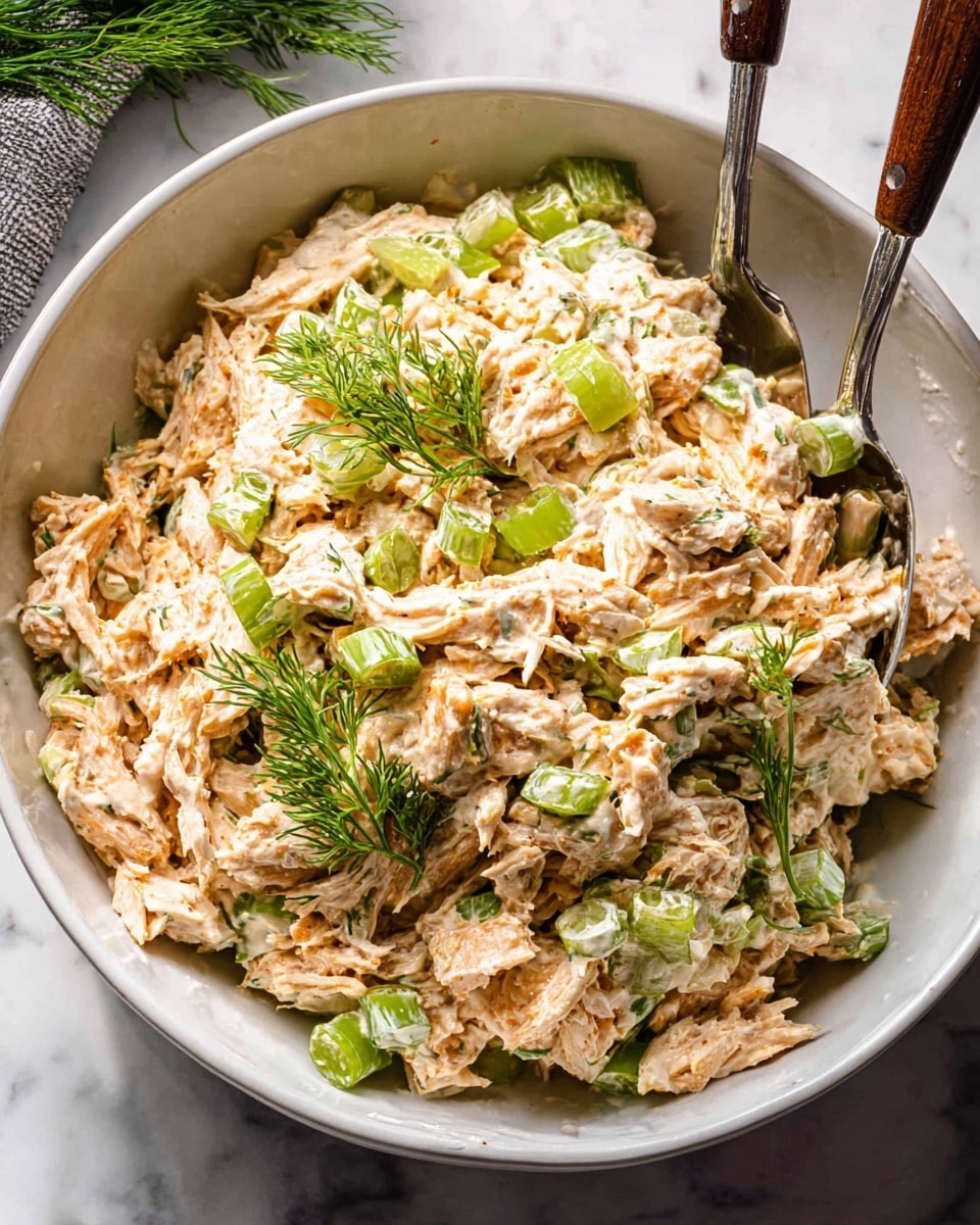 A white bowl filled with a creamy chicken salad showing shredded pale orange chicken mixed with light cream-colored dressing, bright green celery slices, and small green onion pieces. The salad is garnished with fresh green dill sprigs on top. Two metal serving spoons with dark wooden handles rest inside the bowl on a white marbled surface. photo taken with an iphone --ar 4:5 --v 7