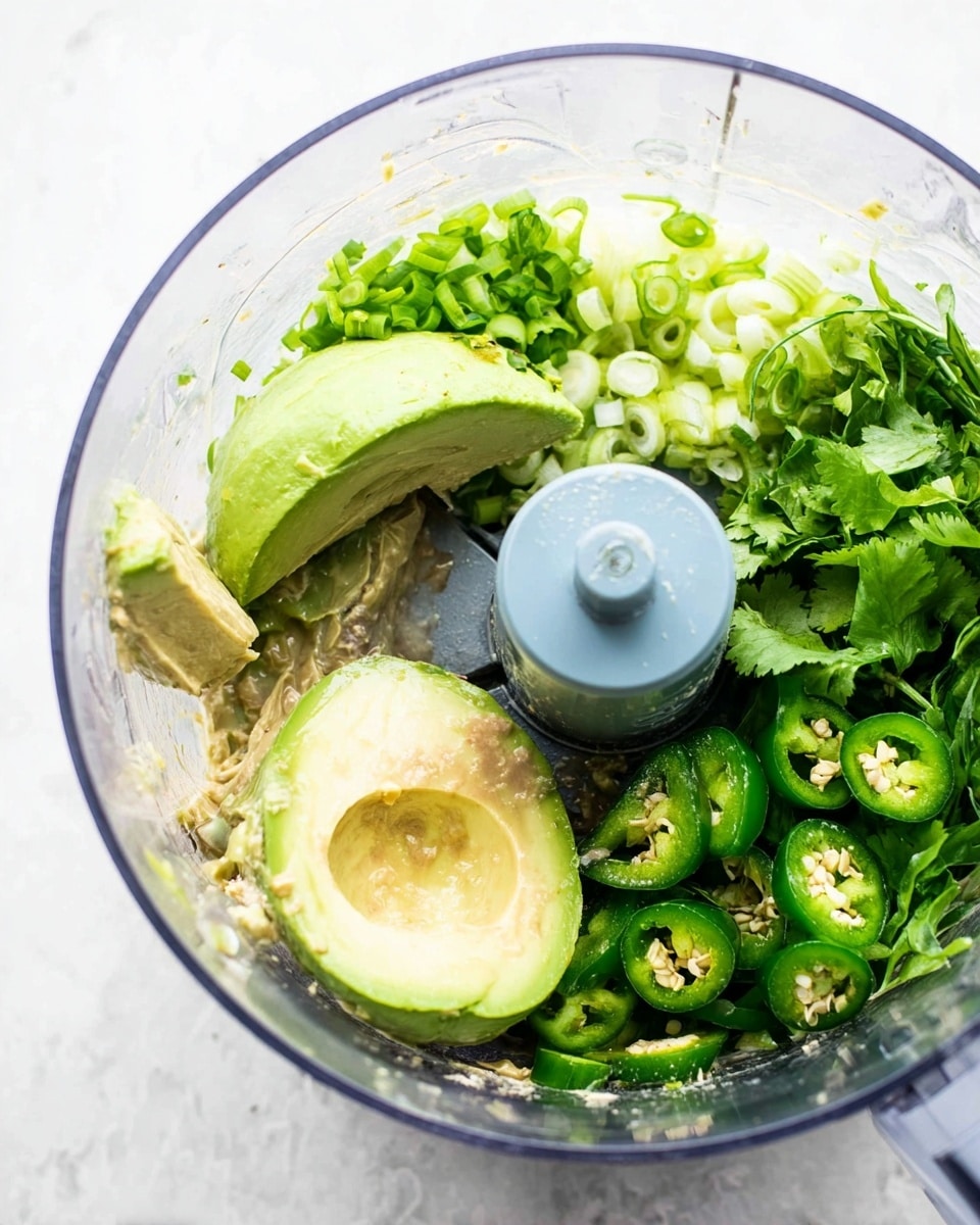 A clear food processor bowl filled with layers of fresh ingredients including large pale green avocado halves in the bottom left, bright green basil leaves behind the avocado, chopped light green scallions next to the basil, sliced dark green jalapeño peppers on the right, and fresh green cilantro leaves near the bottom right. The surface under the bowl is white with a marbled texture. The center blue-gray blade attachment is visible, and some light tan liquid sauce covers part of the avocado. Photo taken with an iphone --ar 4:5 --v 7