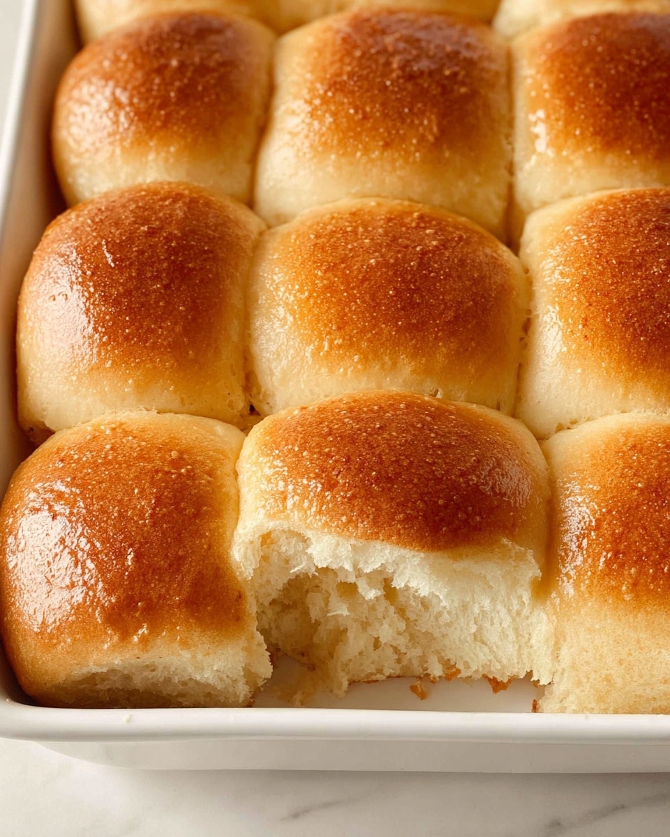 The image shows a close-up of nine golden-brown bread rolls baked closely together in a white rectangular dish. The rolls have a soft, slightly shiny texture on top with a light crust, and one roll in the front has a small piece missing, revealing its fluffy, light interior. The surface beneath the dish is a white marbled texture. The rolls are arranged in a 3x3 grid, filling the dish fully and touching each other. photo taken with an iphone --ar 4:5 --v 7