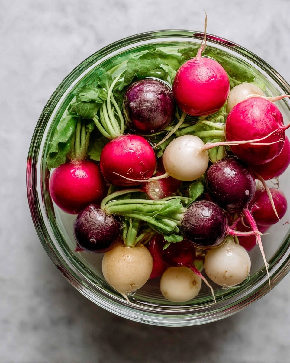 A clear glass bowl filled with a mix of fresh radishes showing three layers: the bottom layer with light beige roots partially under water, the middle layer with round radish bulbs in shades of bright pink, dark purple, and white, and the top layer with green leafy radish stems that look fresh and slightly shiny. The bowl sits on a white marbled surface that adds a clean look. photo taken with an iphone --ar 4:5 --v 7