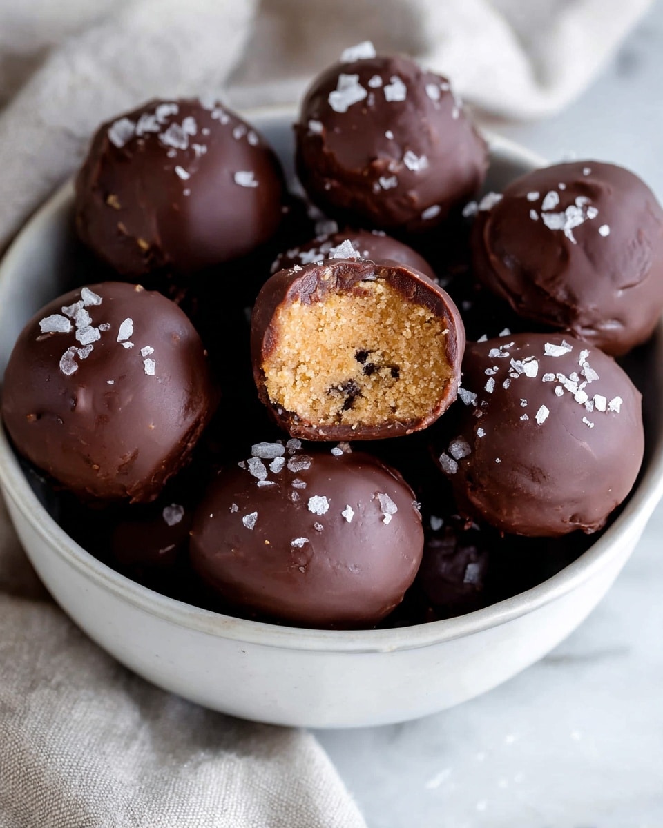 A white bowl filled with round chocolate-covered balls, each topped with small pieces of sea salt. One ball is in the center, showing a bite taken out, revealing a light tan, crumbly inside with small dark bits. The chocolate coating is smooth with some texture and slight shine. The bowl rests on a white marbled surface with a soft cloth nearby. photo taken with an iphone --ar 4:5 --v 7