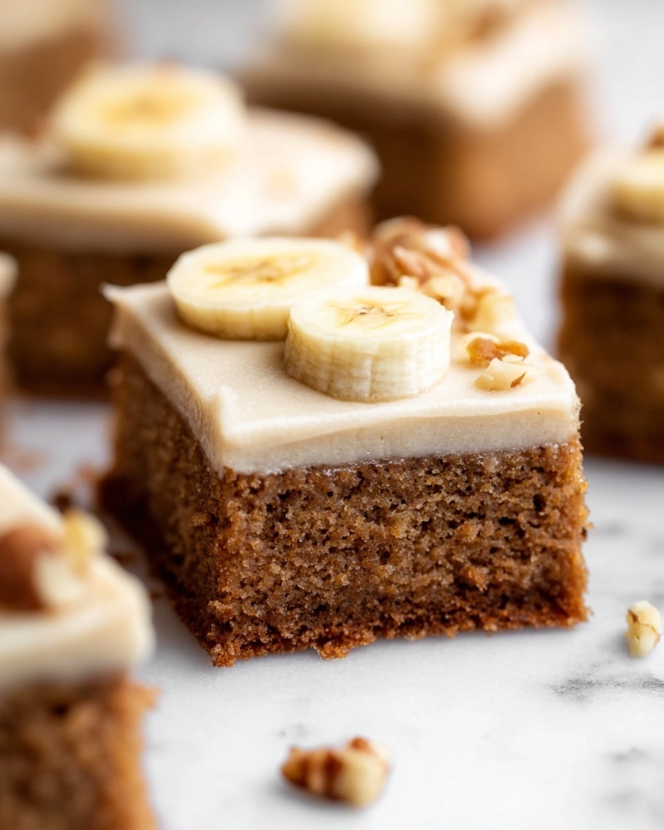 The image shows a close-up of a square slice of cake with two distinct layers on a white marbled surface. The bottom layer is a thick, moist, brown cake with a slightly rough texture. On top, there is a smooth, creamy beige frosting layer that looks soft and thick. Two thin, round slices of banana rest on the frosting, slightly overlapping near the middle. Small pieces of chopped nuts are scattered on and around the cake, adding texture. Other similar slices of the cake are blurred in the background. Photo taken with an iphone --ar 4:5 --v 7