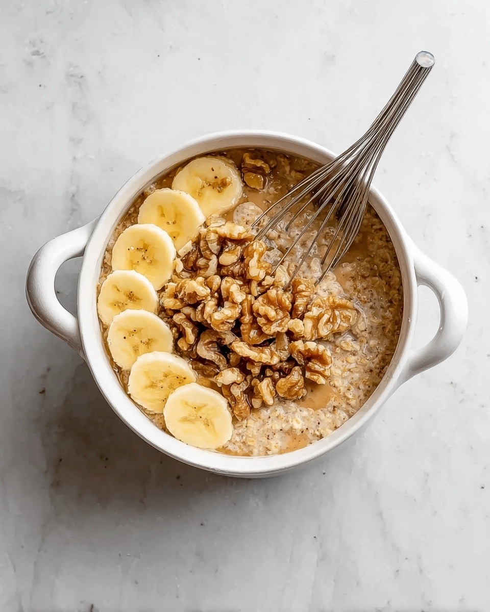 A white bowl with two small handles holds a mixture of oatmeal soaked in a light brown liquid, topped with a cluster of chopped walnuts in the center. To one side of the walnuts, there are several light yellow banana slices arranged in a small pile. A small metal whisk rests inside the bowl, partially submerged in the oatmeal mix with two banana slices balanced on its wires. The bowl is placed on a white marbled surface. photo taken with an iphone --ar 4:5 --v 7
